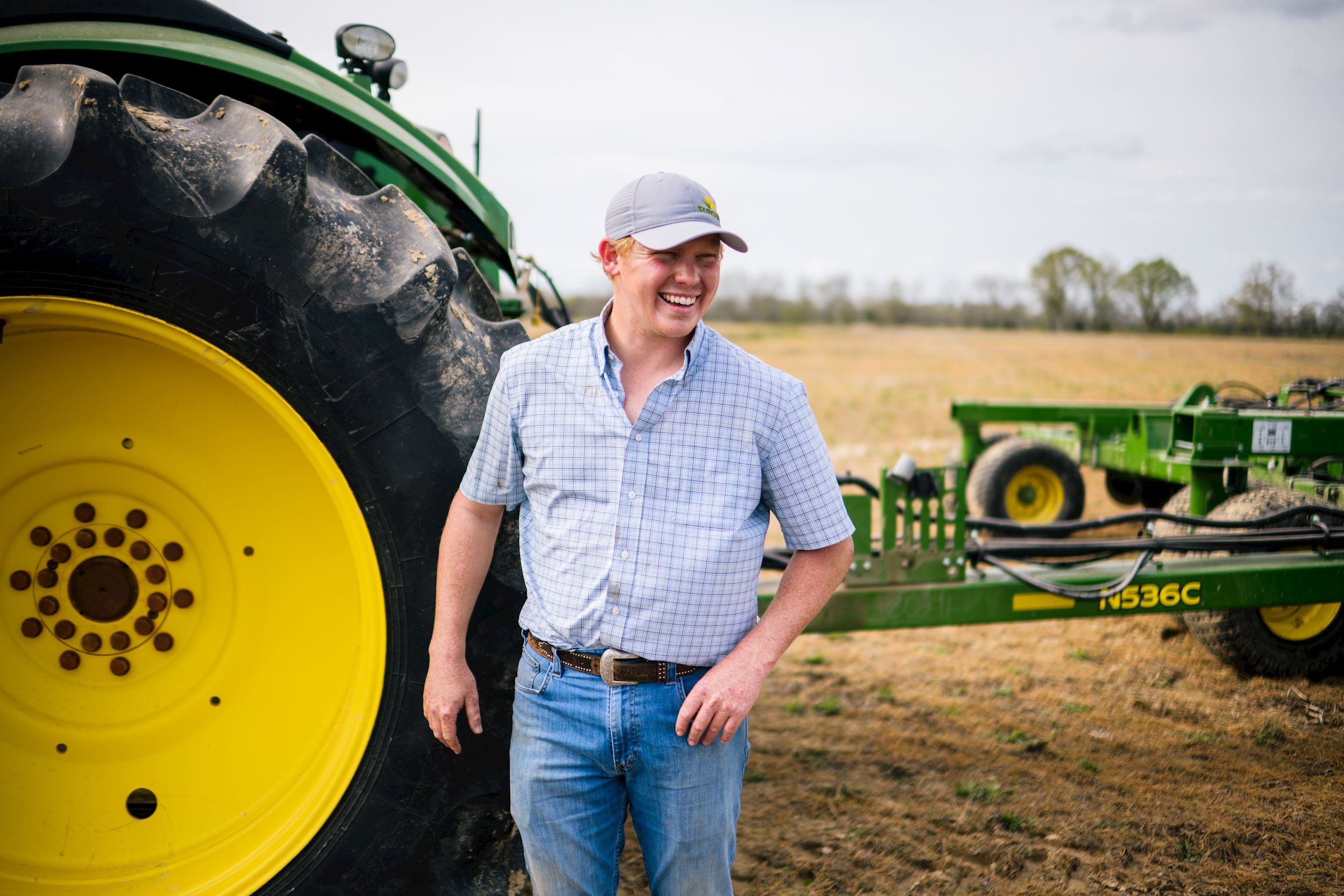 man wearing a hat laughs in front of a John Deere tractor 